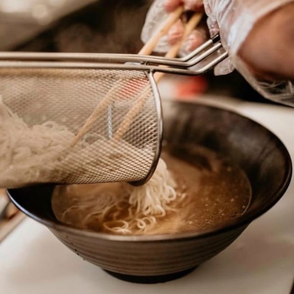 A chef in gloves uses a strainer to add ramen noodles to a bowl of broth.