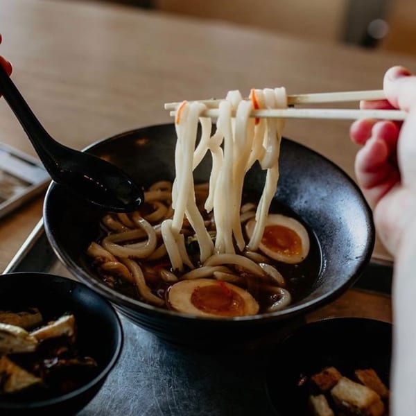 Chopsticks lifting thick ramen noodles from a dark bowl with broth and soft-boiled eggs.