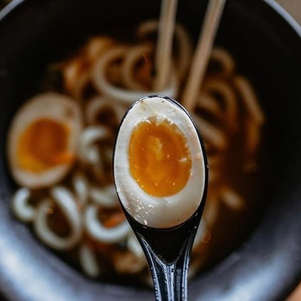 A spoonful of soft-boiled egg with a runny yolk, held over a bowl of Boru ramen.
